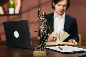 Bronze statue of Lady Justice holding scales in front of a man reading legal documents at a desk with a laptop and books in a modern office setting