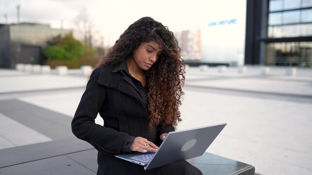 Young professional woman with long curly hair sitting on a bench in a modern city plaza and typing on her laptop, focused on her work