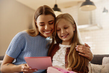 Happy daughter giving greeting card and gift to her smiling mom at home