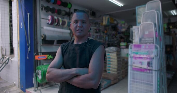 Confident small business hardware store owner standing at shop entrance with arms crossed expressing resilience independence responsibility pride local entrepreneurship