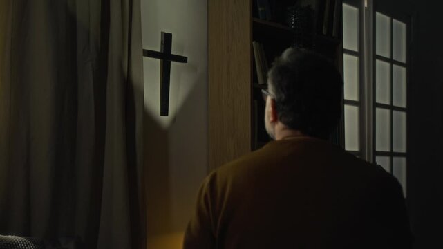 Back view of religious mature man with glasses looking at cross hanging on wall between curtain and bookcase in dimly lit room