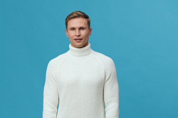 Full body portrait of handsome young man with short light brown hair wearing cozy cream knit turtleneck sweater posing against solid light blue background studio shot. People lifestyle concept.