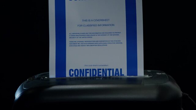 A coversheet to a confidential document is fed into a paper shredder. Subdued lighting. Suggestive of spying, espionage, or cover-up.