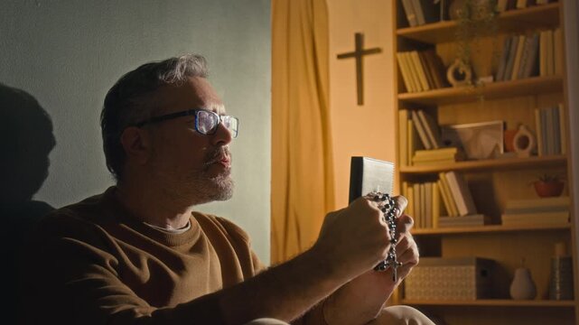 Rack focus shot of Caucasian mature man with glasses praying with eyes closed holding bible and rosary in front of his face and then looking at cross hanging on wall next to bookcase