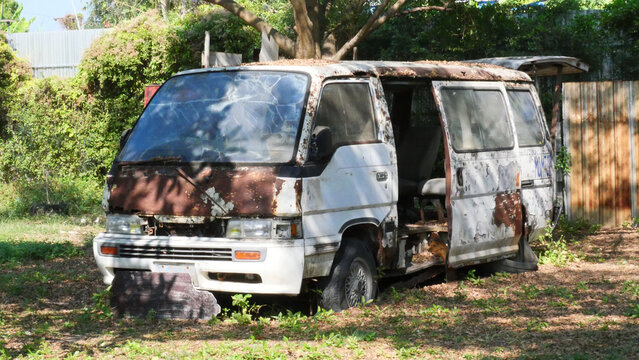 Abandoned Rusted White Van in Overgrown Garden. An old, broken white van with rust and shattered glass sits abandoned in a grassy, overgrown outdoor field.