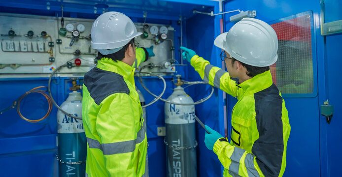 Two Asian engineers inspecting industrial gas manifold system. Professional technicians checking pressure gauges and monitoring oxygen nitrogen argon cylinders at manufacturing plant.