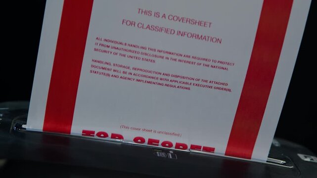 A coversheet to a confidential document is fed into a paper shredder. Subdued lighting. Suggestive of spying, espionage, or cover-up.