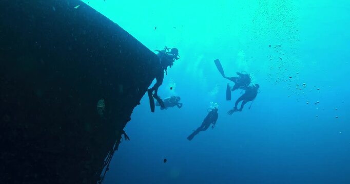 Group of scuba divers explores a massive shipwreck in deep blue water, surrounded by bubbles and soft light rays. Cinematic slow motion underwater adventure and discovery.

