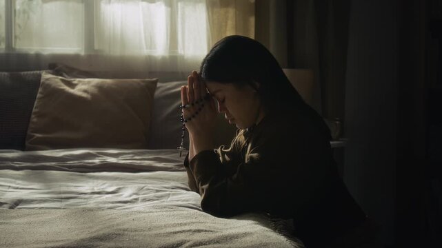 Side view of brunette young woman holding rosary beads praying with hands placed together and bowing her head while leaning on bed with elbows