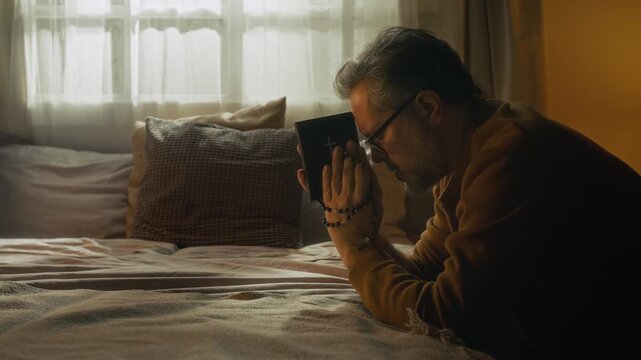 Side shot of mature man with grey hair holding bible book and rosary turning to God during morning prayer while kneeling with elbows on bed