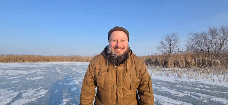 Portrait of a smiling bearded man in winter clothes standing on a frozen lake. Frost on beard in sunny cold weather