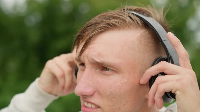 young white man wearing headphones in rainy park, wet hair and hoodie, adjusting earcups and tapping controls while listening intently to music closeup portrait shows undercut hairstyle, raindrops