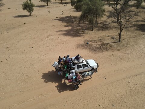 Aerial view of a crowded vehicle overloaded with people and goods traversing a sandy landscape under the harsh sun, Dodji, Louga Region, Senegal.