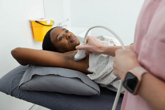 African woman patient lying on exam table, clinician scanning axilla with probe, yellow sharps