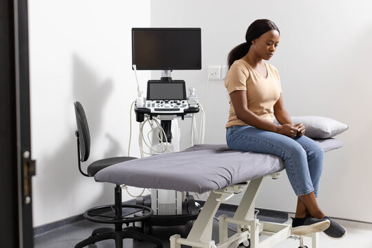 Woman sitting on examination table in clinic next to ultrasound machine and stool, copy space