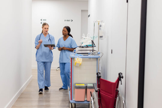 Diverse female nurses walking down hospital corridor in light blue scrubs, one pointing at tablet