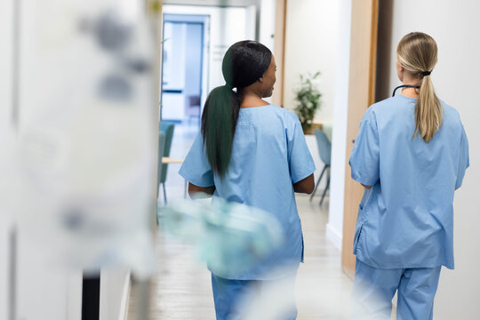 Two female medical workers walking down bright clinic corridor in blue scrubs, one with stethoscope