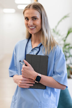 Adult female nurse standing in corridor wearing blue scrubs holding black tablet with stethoscope