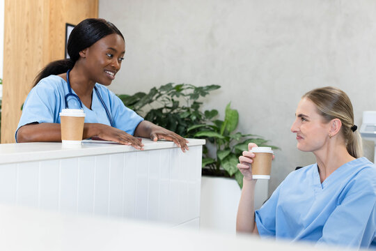 Female healthcare workers in blue scrubs chatting at nurses' station, holding coffee, stethoscope