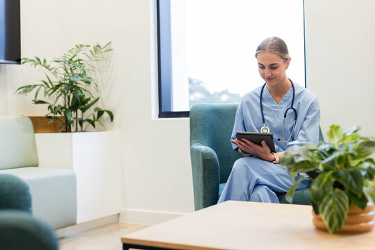 Female nurse sitting on teal armchair near bright window, holding tablet and stethoscope