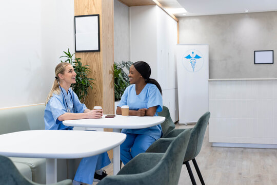 Two female nurses in scrubs chatting while holding coffee at staff lounge with stethoscope nearby