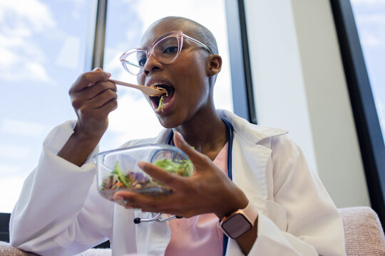 African American woman in lab coat sitting at clinic near windows holding salad bowl eating fork