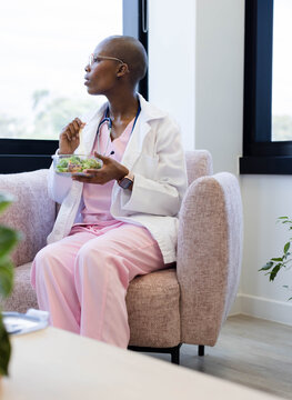 Medical pro wearing pink scrubs is sitting on pink chair by window, holding salad and stethoscope