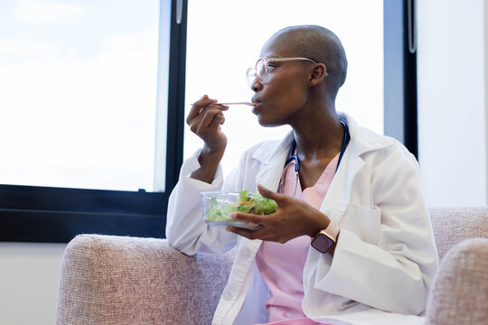Adult African woman sitting on sofa in lounge in lab coat, scrubs, holding salad, fork, stethoscope