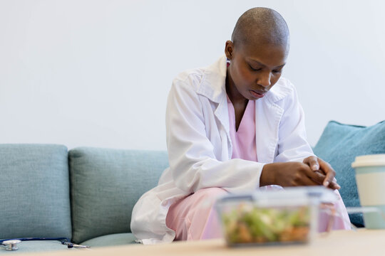 Woman in scrubs, lab coat sitting on teal sofa in clinic with stethoscope, salad container, cup