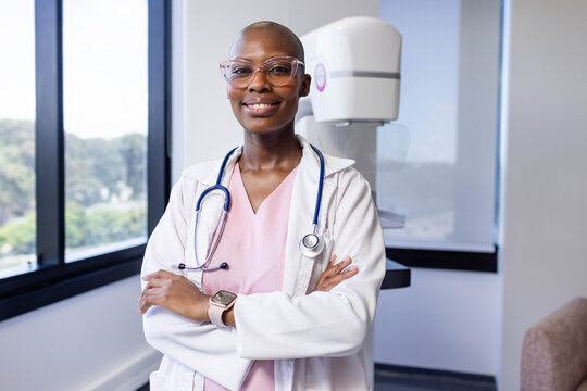 African American woman standing with arms crossed in exam room, wearing lab coat near mammogram