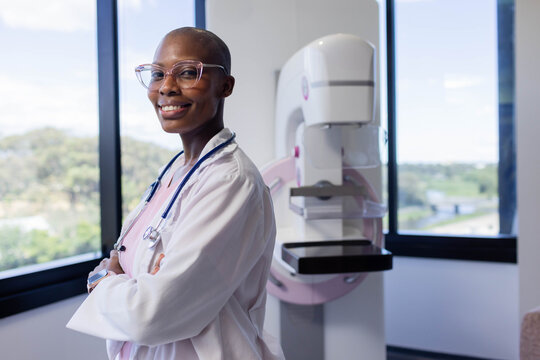 Female doctor standing with arms crossed in radiology room in lab coat and stethoscope by mammogram