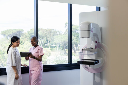 Female radiology tech and patient standing in mammography room reviewing tablet, copy space