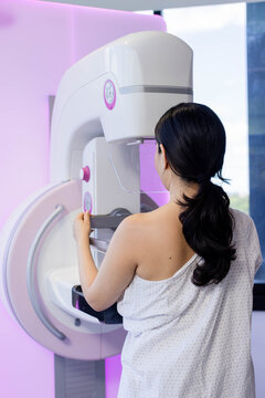Patient is standing with gown off one shoulder, facing mammogram unit with clear paddle near window