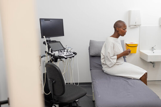 African woman in gown sitting on exam couch holding sharps, US cart showing probes, copy space