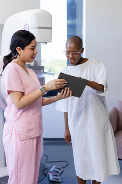 Diverse female clinician in pink scrubs and patient in gown reviewing tablet near mammography unit