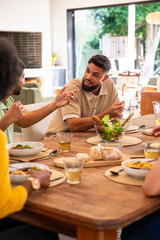 Diverse friends chatting at dining table, Indian man wearing beige polo tossing salad in glass bowl © wavebreak3