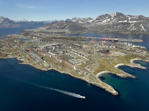 A large Greenland city spreads along a rugged coastline, with modern buildings, industrial areas, and a busy harbor set against dramatic mountains and deep blue Arctic waters under clear skies.