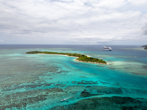 Aerial view of turquoise waters embrace a narrow island fringed with white sand, contrasting with the distant silhouette of a cruise ship on the horizon, Mystery Island, Vanuatu.