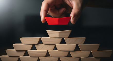 Hand placing a unique red building block atop a wooden pyramid, symbolizing strategic completion, leadership, and the final piece of a successful business structure
