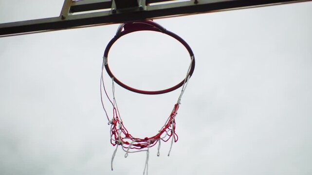 basketball hoop ball suspended midair, frayed red net hanging from rusty rim under overcast sky, ball cutting clean arc toward center, slow cinematic motion captures swish moment and subtle bounce,