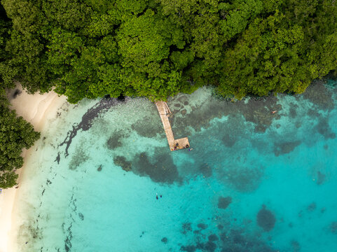 Aerial view of a wooden pier stretching into the turquoise sea, fringed by a vibrant green forest and a pristine white sandy beach, Espirito Santo, Sanma Province, Vanuatu.