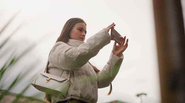 young woman taking phone selfie outdoors in urban setting wearing puffer jacket and white handbag, braided hair, candid pose capturing social media content with shallow depth of field, overcast sky,