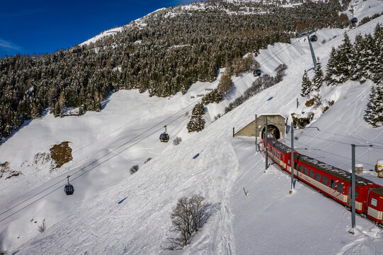 Aerial view of a vibrant red train emerges from a tunnel amidst the snow-covered mountains, with cable cars ascending towards the peaks, Andermatt, Switzerland.