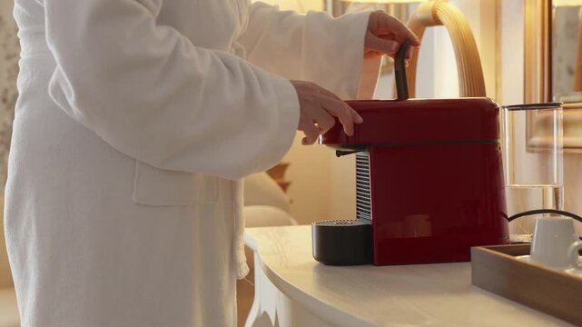 Adult person in white bathrobe placing coffee capsule and cup into red coffee machine on kitchen counter, cozy morning routine and home comfort concept.