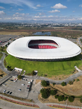 Zapopan, Mexico - December 7 2025: Aerial drone view of modern Akron Stadium in Guadalajara. White roof, red seats, green surroundings, parking, and distant city under blue sky with clouds