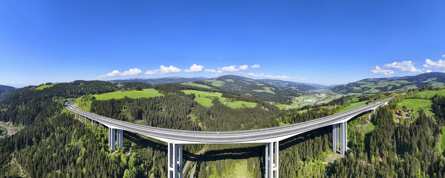 Aerial view of a modern bridge soaring through the Lavanttal valley, contrasting with the lush green forests and verdant fields under a clear blue sky, Wolfsberg, Carinthia, Austria.