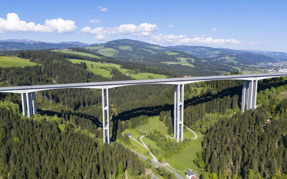 Aerial view of a long concrete bridge soaring through a vibrant green valley, connecting distant hills under a clear blue sky, Wolfsberg, Carinthia, Austria.