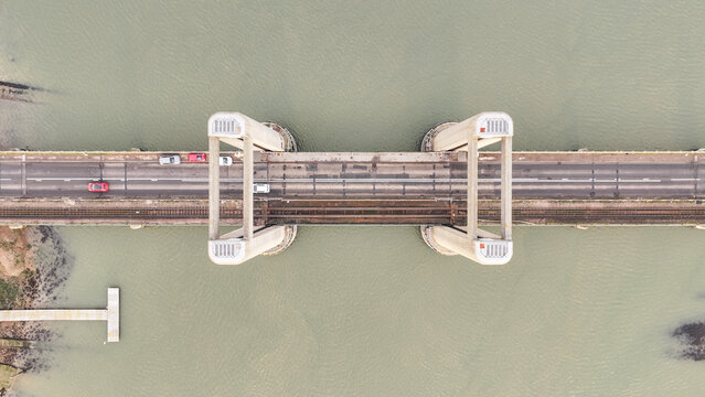 Aerial view of cars traverse the Kingsferry Bridge, its concrete pillars stark against the Swale's muted waters, a modern artery amidst the serene landscape, Sittingbourne, United Kingdom.