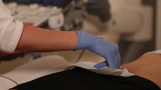 A medical professional wearing blue latex gloves performs an ultrasound examination on a patient, carefully moving the transducer probe over a protective paper towel on the lower abdomen.

