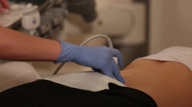 A medical professional wearing blue latex gloves performs an ultrasound examination on a patient, carefully moving the transducer probe over a protective paper towel on the lower abdomen.

